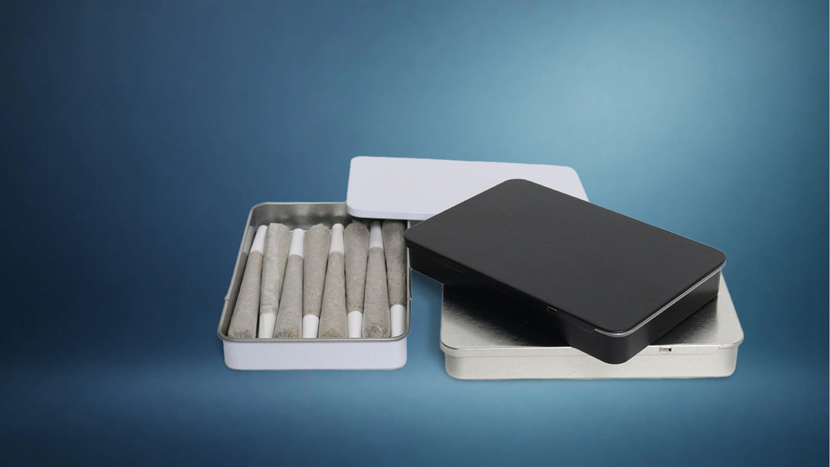Black, white, and silver metal cannabis pre-roll slider tins with pre-rolls on a blue gradient background.